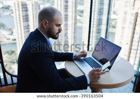 Successful man managing director is reading message on mobile phone during work on portable net-book. Skilled male entrepreneur is using cell telephone, while is sitting with laptop computer in office