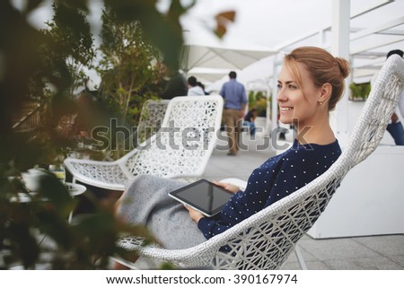 Young cheerful female student is enjoying free time,while is sitting with touch pad in coffee shop outdoors. Beautiful woman is holding digital tablet and smiling for someone during rest in cafe-bar