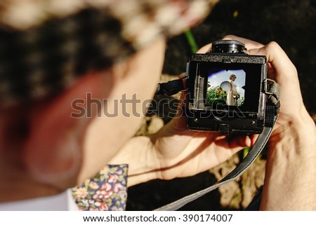 stylish handsome groom holding medium format film camera, taking photo of gorgeous bride in sunny sunflower field