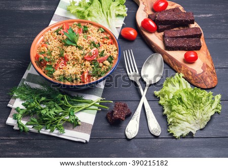 Couscous in a blue ceramic bowl, parsley, dill, toasted bread croutons, silver fork and spoon, cherry tomatoes, lettuce leaves on a dark black wooden background