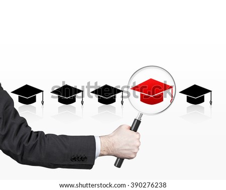 Hand holding magnifier over red picture of academic hat in row of black hats. White background. Concept of choice.