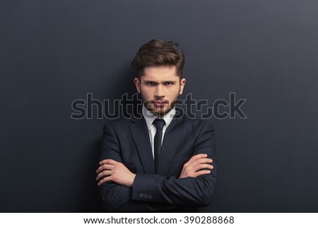 Handsome young student in classic suit is looking at camera, standing with crossed arms against blackboard