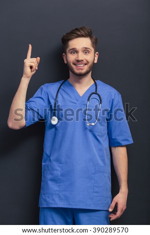 Handsome young doctor in blue medical uniform is pointing up, smiling and looking at camera, standing against blackboard