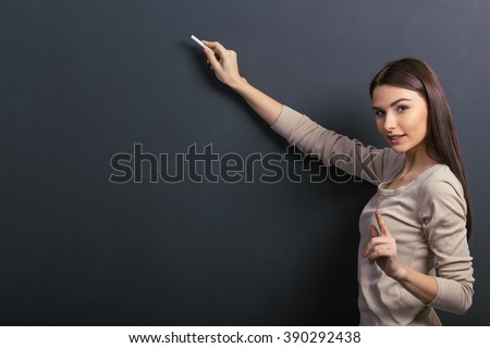 Beautiful young female student is holding a piece of chalk and looking at camera, standing against blackboard