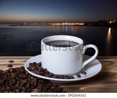 Coffee beans and coffee in white cup on wooden table opposite a defocused natural landscape for background. Collage. Selective focus.