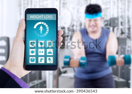 Hand holding smartphone with weight loss monitoring application on the screen, shot with overweight person doing workout at gym