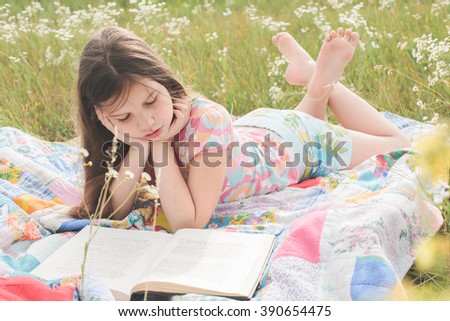 Little girl lies on the patchwork rug on the field, eats a candy and reads a book in the light sunny summer day