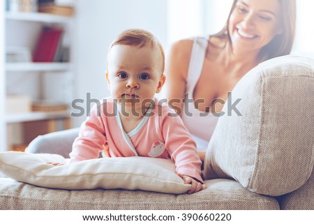 Cutie with mommy. Little baby girl looking at camera and leaning on cushion while her mother sitting on the couch at background