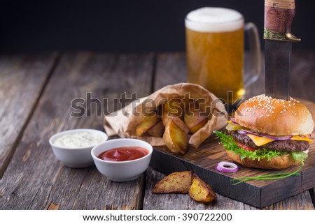 Appetizing burger with beef patty, lettuce, tomato, cheese, pickled cucumbers and red onion served with fried potato, white sauce and ketchup, on wooden cutting board. With mug of light beer