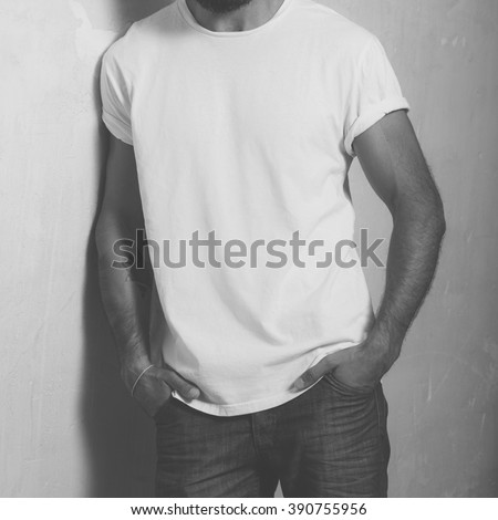 Closeup photo bearded stylish man with tattoo wearing white blank t-shirt and black jeans, standing opposite concrete empty wall. Vertical ,mockup