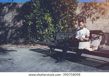 Photo bearded man wearing white tshirt sitting city park bench and reading book. Studying at the University, working new project. Books, laptop, backpack bench. Horizontal