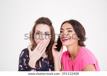 Shocked beautiful young woman overhearing conversation of cheerful female with mobile phone over white background