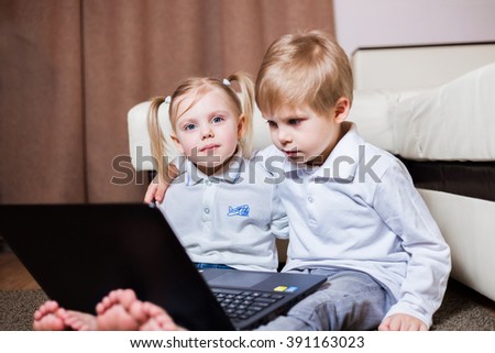 blond little children girl and boy in jeans sitting barefoot on the floor with a laptop watching cartoons, horizontal frame