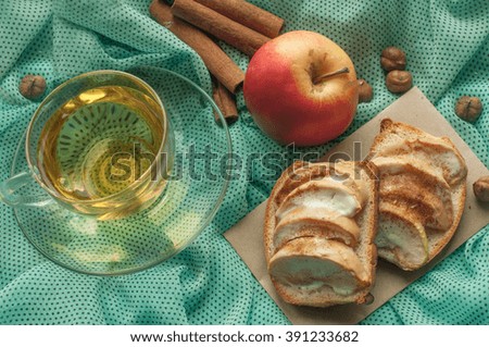 A photo with a cup of herbal tea, toasts with apple slices and cream cheese, fresh apples, nuts and cinnamon sticks  