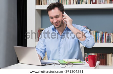 Handsome male freelancer working on laptop computer in library and speaking over smart phone with business partners.