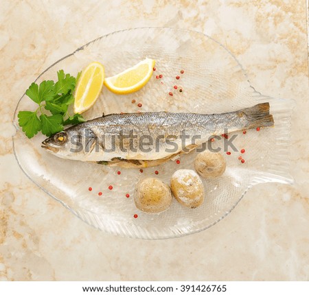 marine fish seabass. cooked in the oven. on a transparent plate in the shape of a fish. with baked potatoes, lemon, parsley and spices. top view
