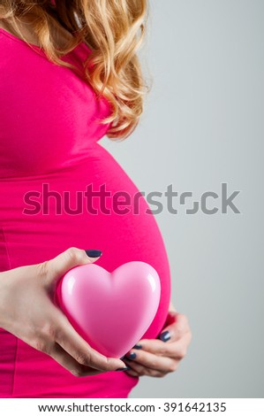 Young beautiful pregnant girl in pink t-shirt hugs her belly with her hand and holds heart, vertical image