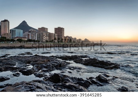 Cape Town Seapoint promenade with city buildings, Lion's Head mountain peak in the background and ocean waves in the foreground at sunset. Royalty-Free Stock Photo #391645285