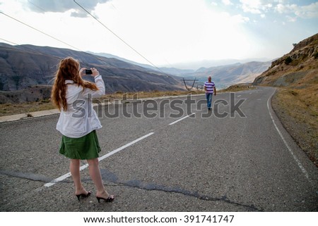 Young woman with red hair taking pictures in the camera man her husband, walking on the asphalt road between the mountains