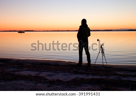 Silhouette of tall nature photographer at tripod taking picture on the beach at sunset time 