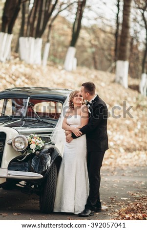 Stylish wedding couple, bride, groom kissing and hugging near retro car in autumn