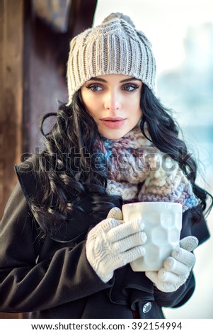 Beautiful girl is drinking a coffee from big white cup. Outside photo shoot. Winter. A girl is in warm clothes and with hat on head. Nice make up and curly hairs.
