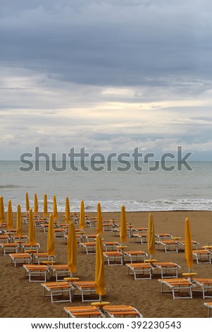 Sea beach with shut yellow sun umbrellas and orange chaise lounges standing in line on beige sand against cloudy sky murky day bad weather on seascape background, vertical picture