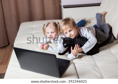 two happy little baby boy and a fair-skinned blonde girl (brother and sister) lie on his stomach on a white sofa with a laptop and watching cartoons on the computer young black stroking a cat