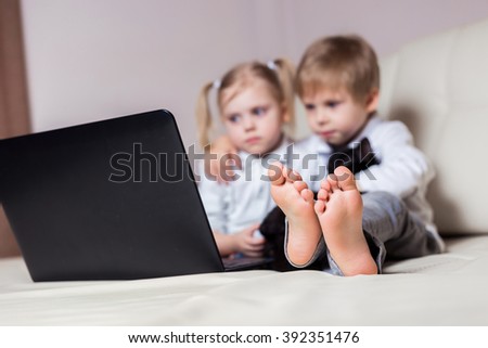 two happy young children: white-skinned boy blond girl sitting with a black cat and a laptop (brother and sister) on a white sofa watching cartoons in the computer, people blurred, legs in Focus