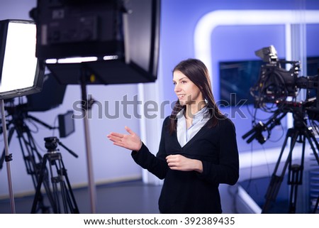 Television presenter recording in television news studio.Female journalist anchor presenting business report.News camera,light equipment behind the scenes.Talking at camera to the TV audience.
