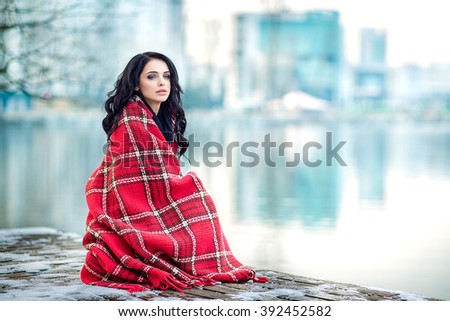 Beautiful woman outdoors is sitting on pier with red plaid. Winter. Young model girl enjoying weather. Fashion model. Nice makeup and hairstyle. 