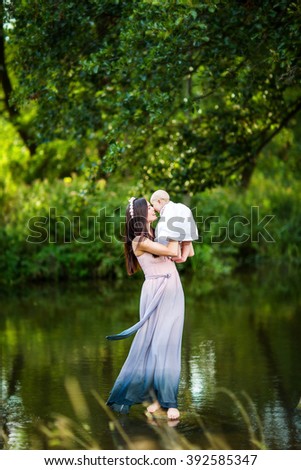 beautiful woman and mother with long  brunette hair in long dress standing in the water of the river or lake on the nature and holding her baby on the hands hugging and kissing her
