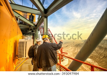 Two coal mine engineers with protective helmets standing and talking on a huge drill machine, talking and watching at the digging site. Beautiful and colorful sky in the background. Rear view. Royalty-Free Stock Photo #393097984