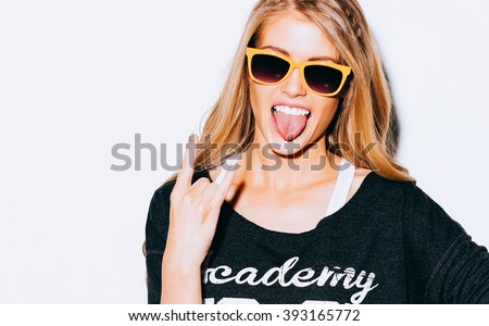 Lovely young blond woman making a call me gesture and showing tongue in suglasses, Sweatshirt and white shorts. White background. Close up. Indoor. Warm color. Hipster.