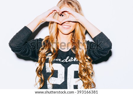 Love. Closeup portrait smiling happy young woman with long blond hair, making heart sign, symbol with hands white wall background. Indoor. Warm color. Hipster.