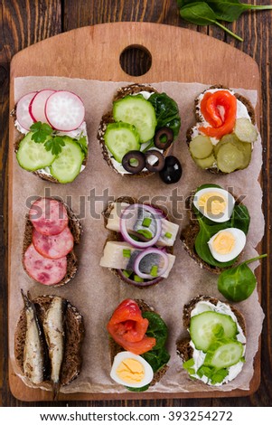 Small sandwiches with vegetables, fish and sausage on board on wooden background. Top view.