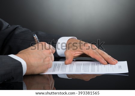 Close-up Of Businessperson Hand Signing Contract Paper With Pen At Desk