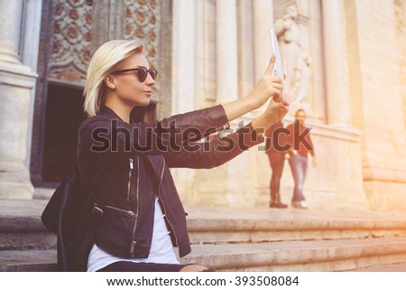 Young hipster girl with trendy look is shooting video on portable touch pad of a street view during her travel abroad. Charming woman is photographing herself with front camera of digital tablet 