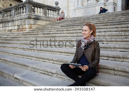 Happy female is smiling for someone, while is sitting with digital tablet in hands on a building stairs outdoors. Female student with good mood is holding touch pad, while is resting after lectures