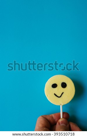 Hand holding smiling yellow lollipop on blue paper. Fools day birthday background. Space for text, lettering, copy.