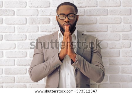 Handsome Afro American man in classic suit and glasses is keeping palms together like praying, standing against white brick wall