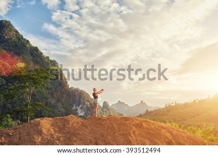 Woman tourist is taking photo of the sunset with mobile phone camera, while is standing against jungle landscape background with copy space for your advertising text message or promotional content