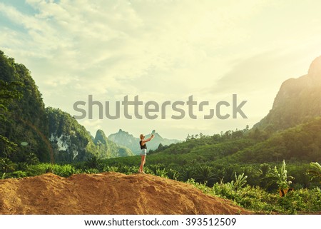 Hipster girl wanderlust is taking photo with mobile phone camera during unforgettable travel in Thailand. Young female is shooting video on cell telephone, while is standing against Amazon scenery