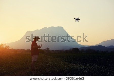 Young man traveler is shooting video on drone, while is standing in rural against high mountains in evening. Male wanderer is using radio-controlled quad-copter during his summer weekend abroad