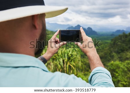 Back view of bearded man is taking photo with cell telephone camera, while is standing on mountain with subtropical forest. Young hipster guy is shooting video on smart phone of a amazing wild nature