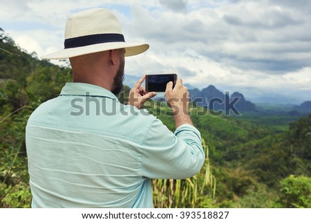 Back view of a young man traveler is taking photo with cell telephone camera of a beautiful jungle landscape. Young male wanderer is shooting video on mobile phone during summer adventure in Asia 