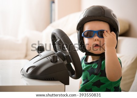 Cute boy toddler sitting at a table in the room and playing with a computer steering wheel. Trainer for a novice driver. Computer Driving Simulator. Early development, home games Royalty-Free Stock Photo #393550831