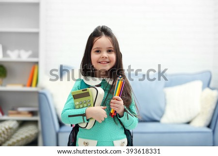 Little girl with green back pack holding stationery and calculator in living room Royalty-Free Stock Photo #393618331