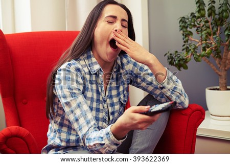 yawning young woman sitting on the red chair and holding tv remote control