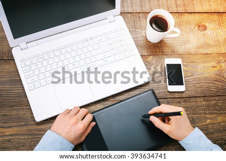 Top view creative photo of objects and hands on vintage brown wooden table. There are mobile phone, coffee, graphics tablet and laptop on it with space for your logo